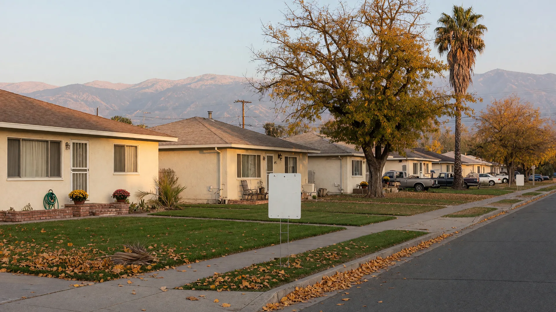 A quiet single-family residential street in Merced County on a clear late-October morning, single-story cream stucco ranch homes with green lawns, valley oak and palm trees, golden autumn leaves on the sidewalk, and the hazy Sierra Nevada foothills on the horizon