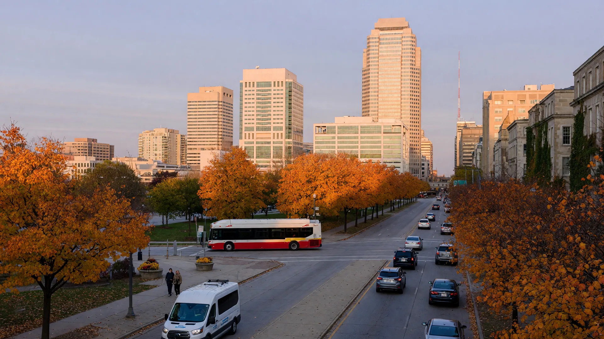 An early October morning commute on Euclid Avenue in Cleveland heading toward University Circle, with the Cleveland Clinic main campus buildings rising in warm low-angle sunlight, an RTA HealthLine bus in the middle distance, and maple trees turning orange and gold