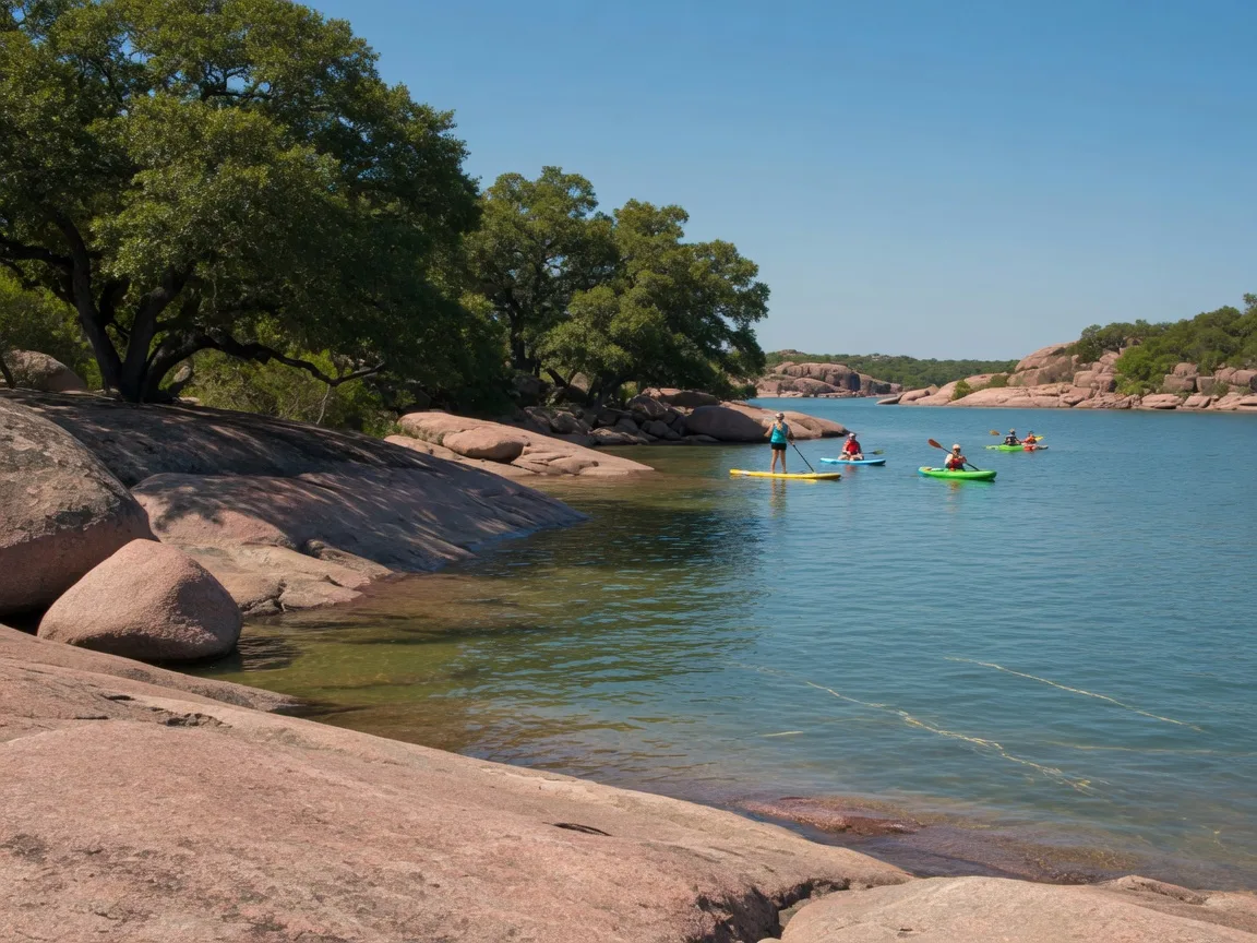 Smooth pink-granite boulders and rock shelves along the clear blue-green water of Inks Lake State Park near Burnet, with paddleboarders and kayakers in the middle distance and live oaks at the waterline on a sunny afternoon