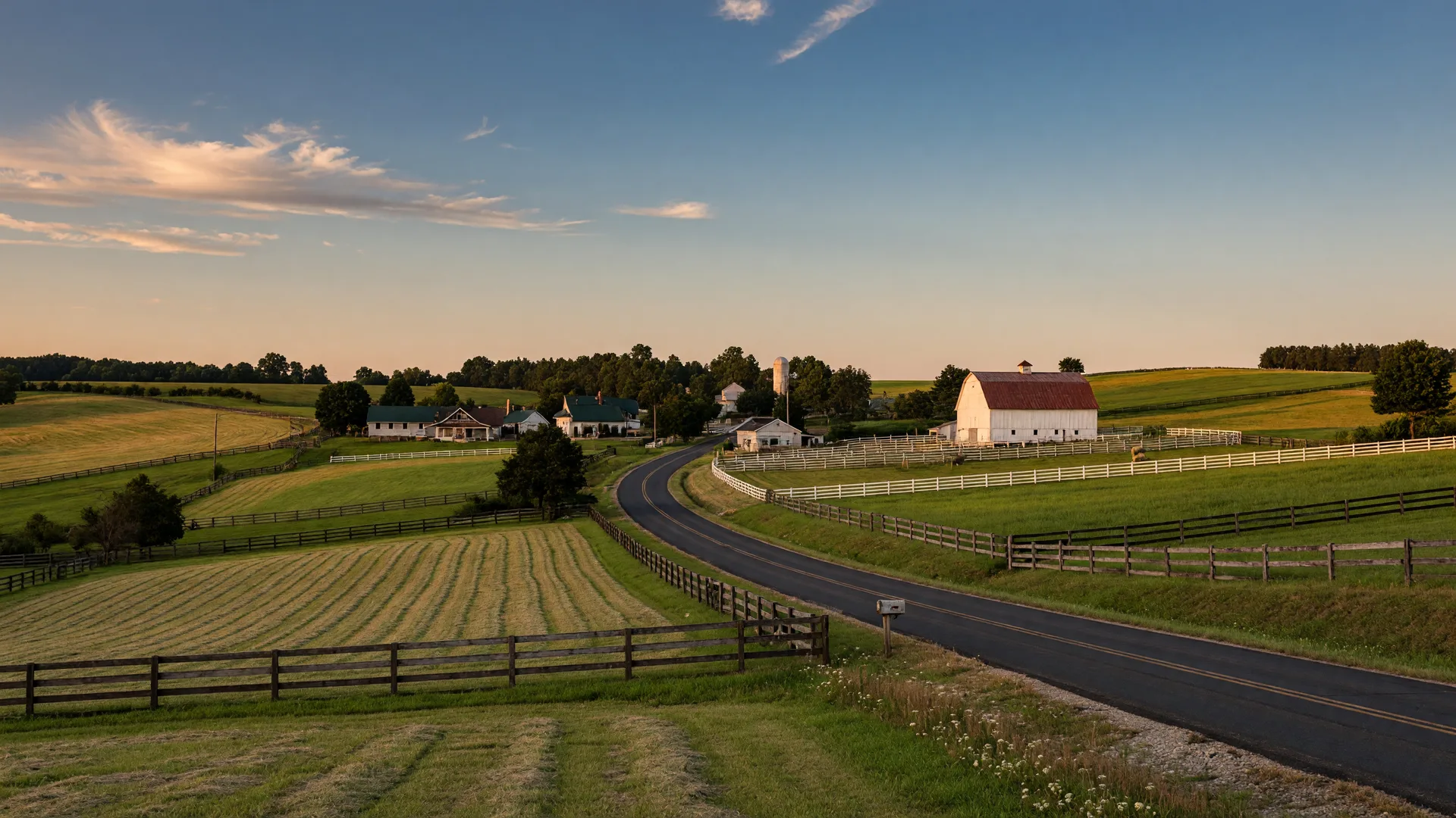 Rolling green Warren County farmland with tree lines along fences and a two-lane road curving toward rural rooftops at golden hour