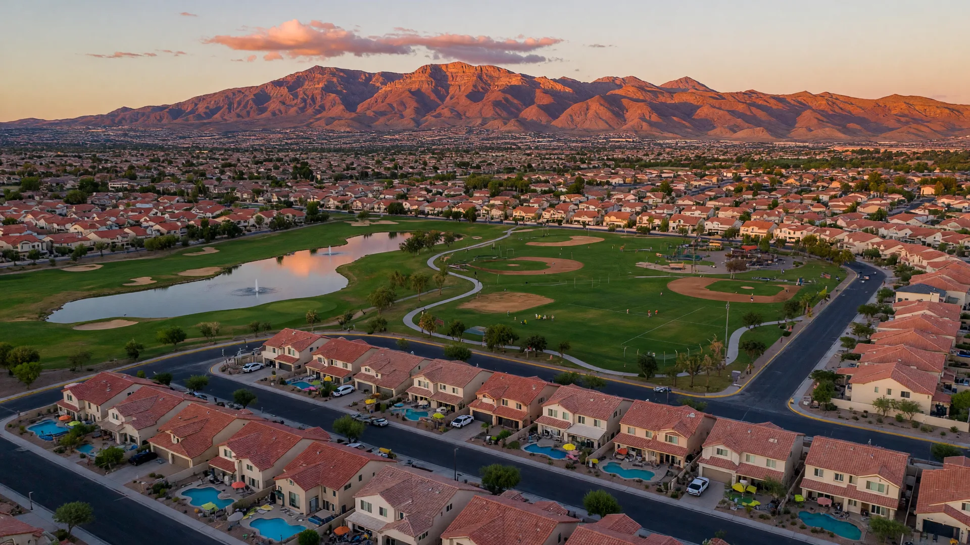 Golden-hour aerial of Southwest Las Vegas master-planned communities in October with red-tile roofs, a regional park, a golf course, and the Spring Mountains