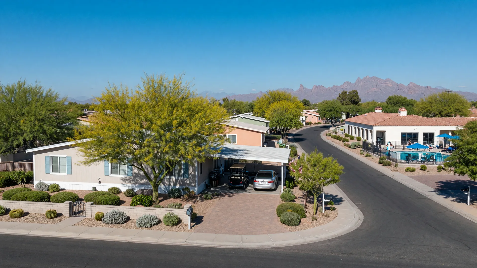 A single-story manufactured home in a 55+ community in Mesa, Arizona on a clear October morning with desert landscaping, a paver driveway, a clubhouse and pool down the street, and the Superstition Mountains on the horizon