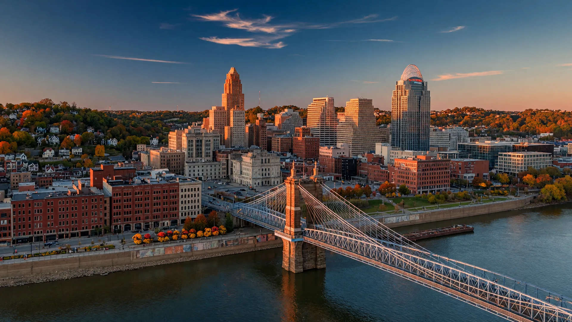 Cincinnati, Ohio at golden hour in early autumn: the Ohio River and Roebling Suspension Bridge in the foreground, the downtown skyline beyond, and hillside neighborhoods with trees turning orange and gold