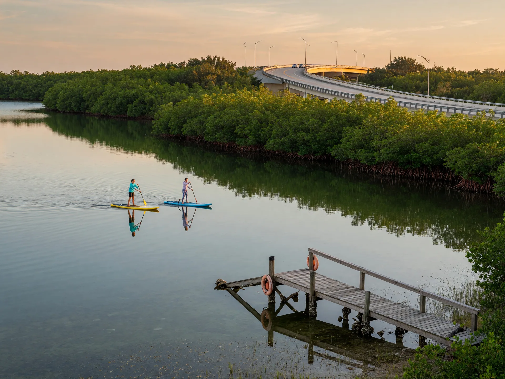 Paddleboarders gliding across the calm, shallow water of Palma Sola Bay at the causeway park, with a small dock in the foreground and the causeway bridge in the distance