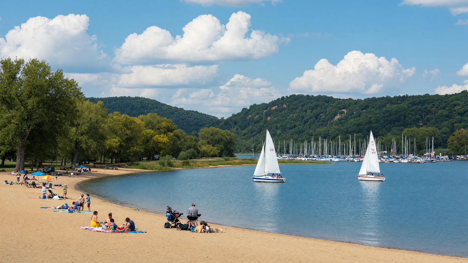 A sweeping golden sand beach and calm blue water at Buck Creek State Park in Springfield Ohio, with sailboats, a distant marina, and wooded green hills under a bright late-summer sky