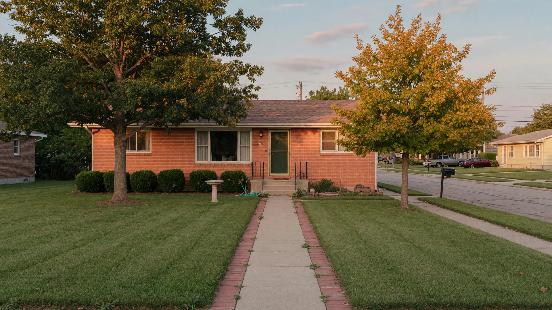 Single-story brick ranch home with a neat walkway and mature shade trees in soft late-afternoon light