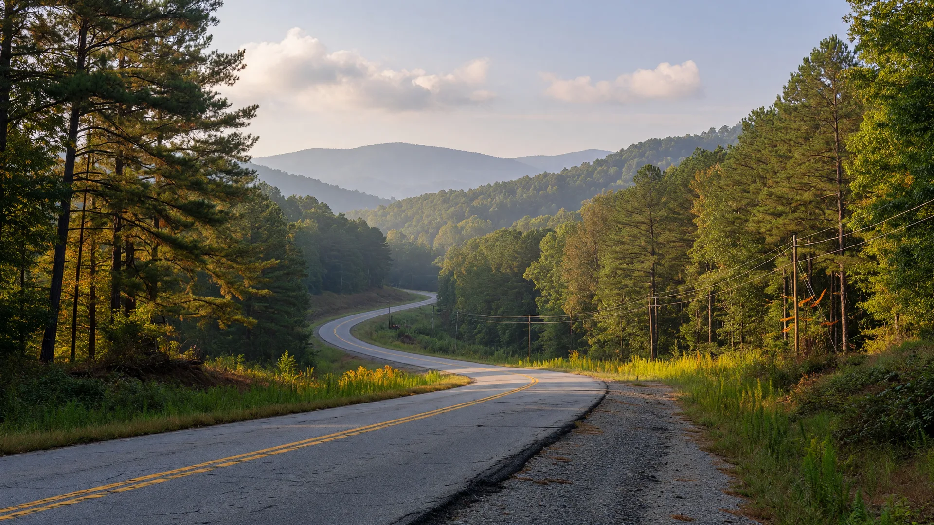 Two-lane rural highway winding through timbered North Georgia foothills on a clear late-summer morning