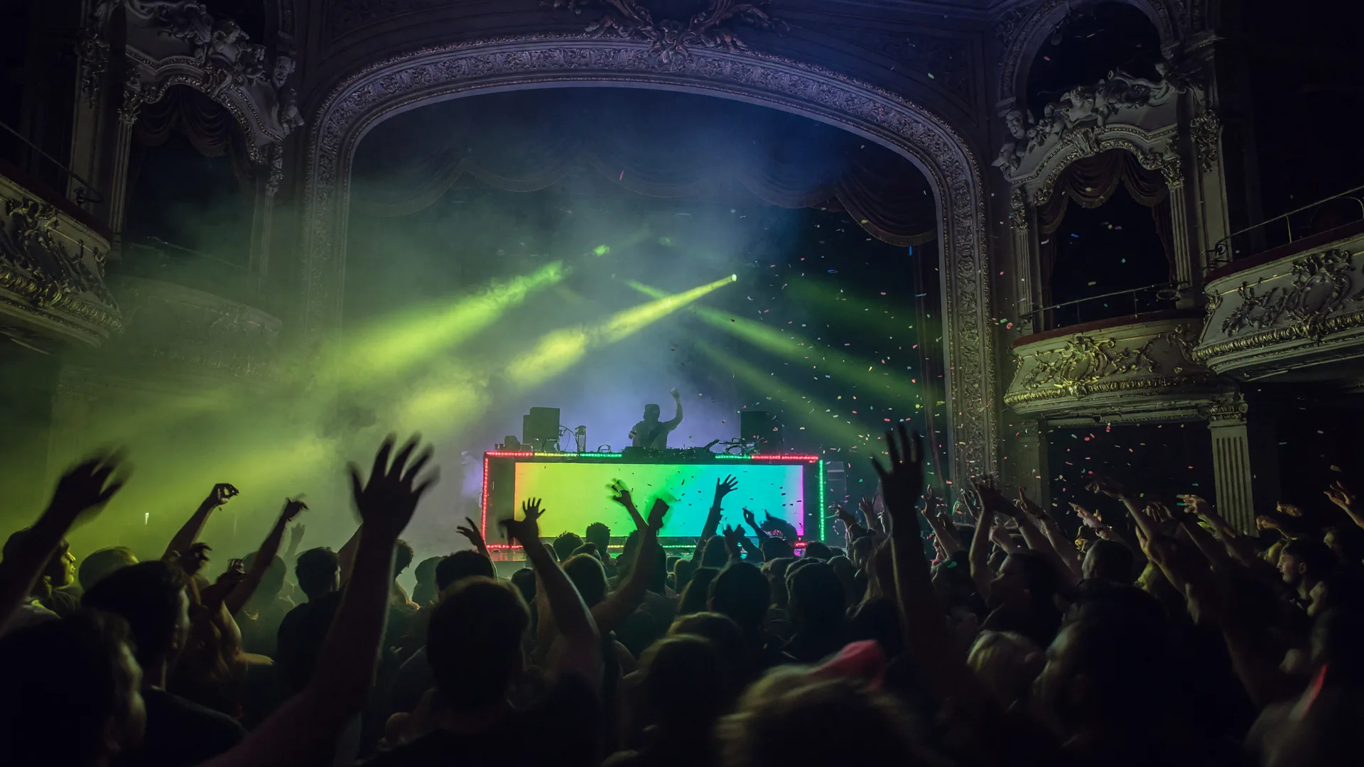A packed crowd dancing under swirling green rave lights inside a historic theater with fog and confetti