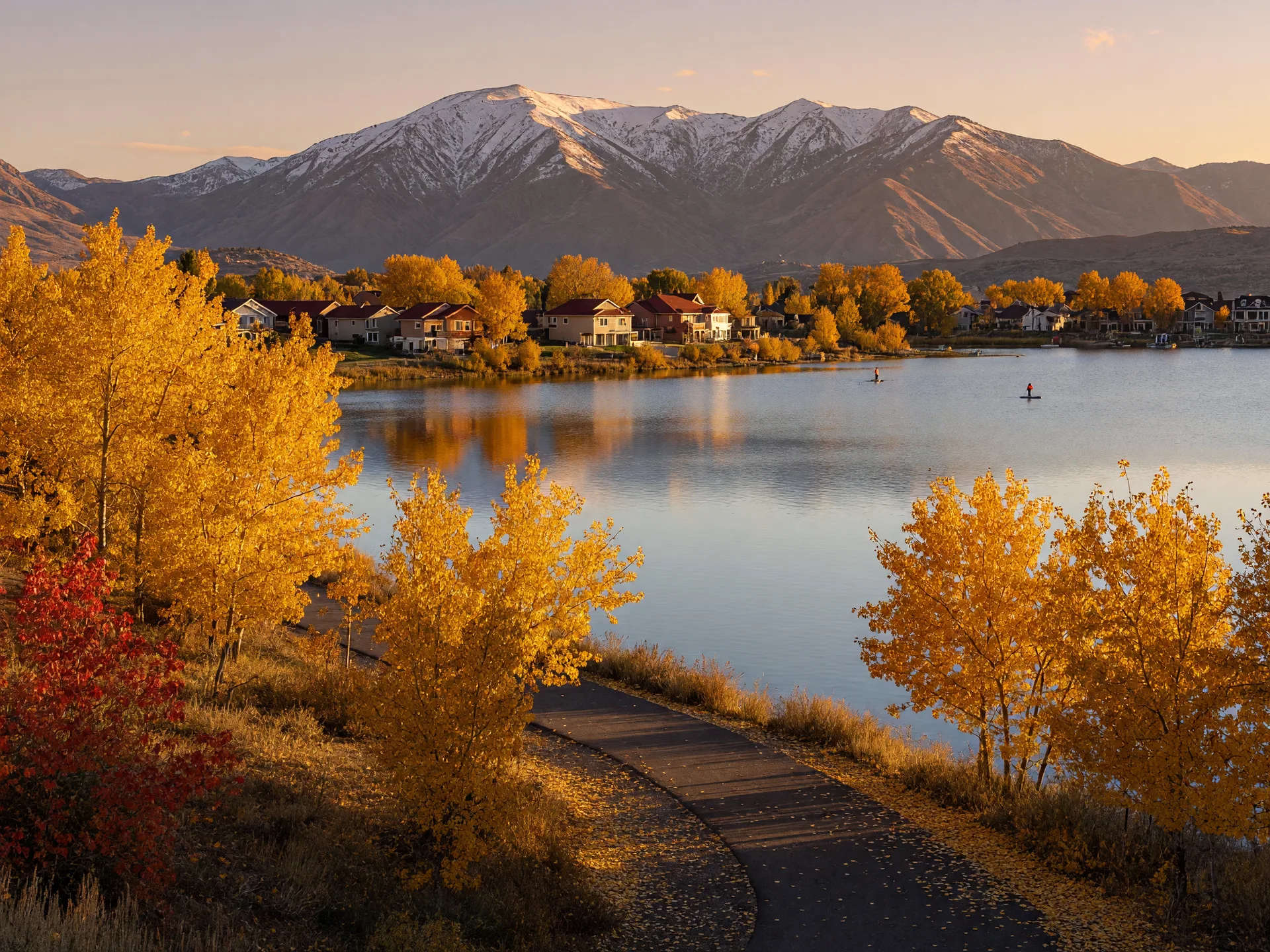 The tree-lined shore of Oquirrh Lake in Daybreak South Jordan Utah in late October with cottonwoods and maples in gold and amber above a calm reflective lake and the snow-dusted Oquirrh Mountains behind