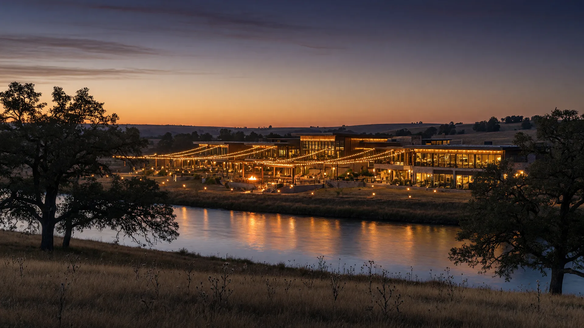 A modern entertainment resort glowing with warm lights across the Red River valley at dusk, seen from the Northeast Texas side, with oak trees and pasture in the foreground