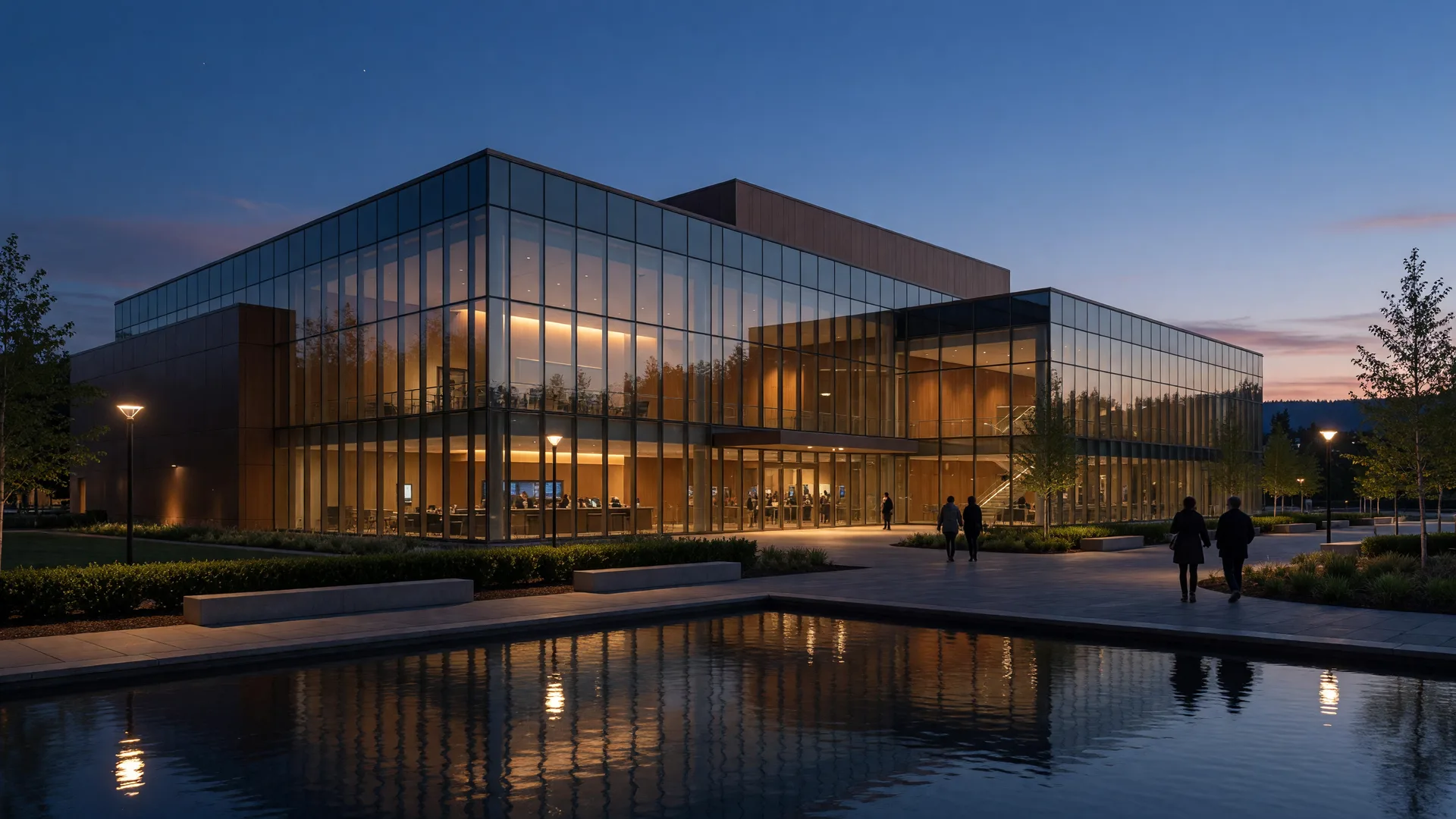 A modern performing arts center glowing with warm amber light at dusk, with a landscaped plaza and reflecting pool in front