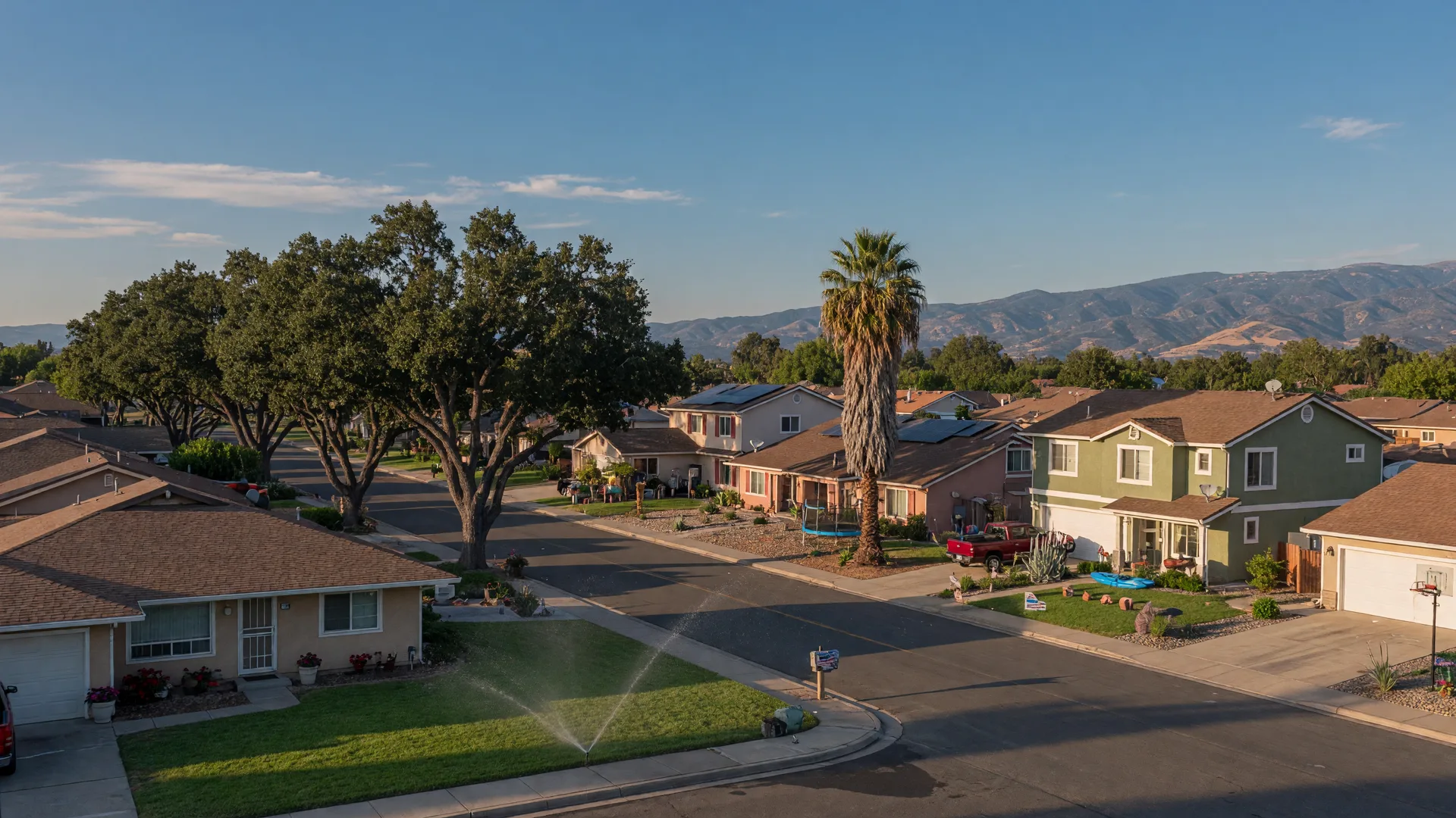 Residential street in Merced, California on a late-summer afternoon with ranch-style and tract homes, manicured lawns, oak and palm trees, and the Sierra Nevada foothills on the horizon