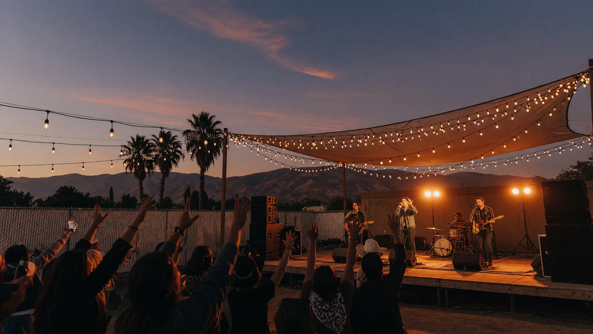 An outdoor evening concert at a Central Valley venue in early October, a wooden stage glowing with warm amber lights and string lights, a silhouetted crowd with raised hands, palm trees and the Sierra Nevada foothills on the horizon at dusk