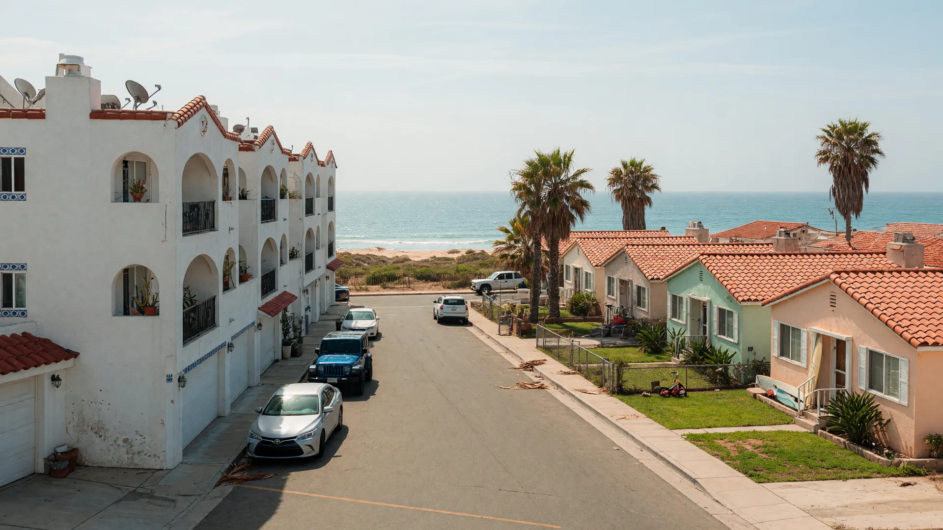 A sunlit Imperial Beach coastal street in ZIP 91932 with a two-story stucco condo building on one side and single-family beach bungalows with tile roofs and palm trees on the other