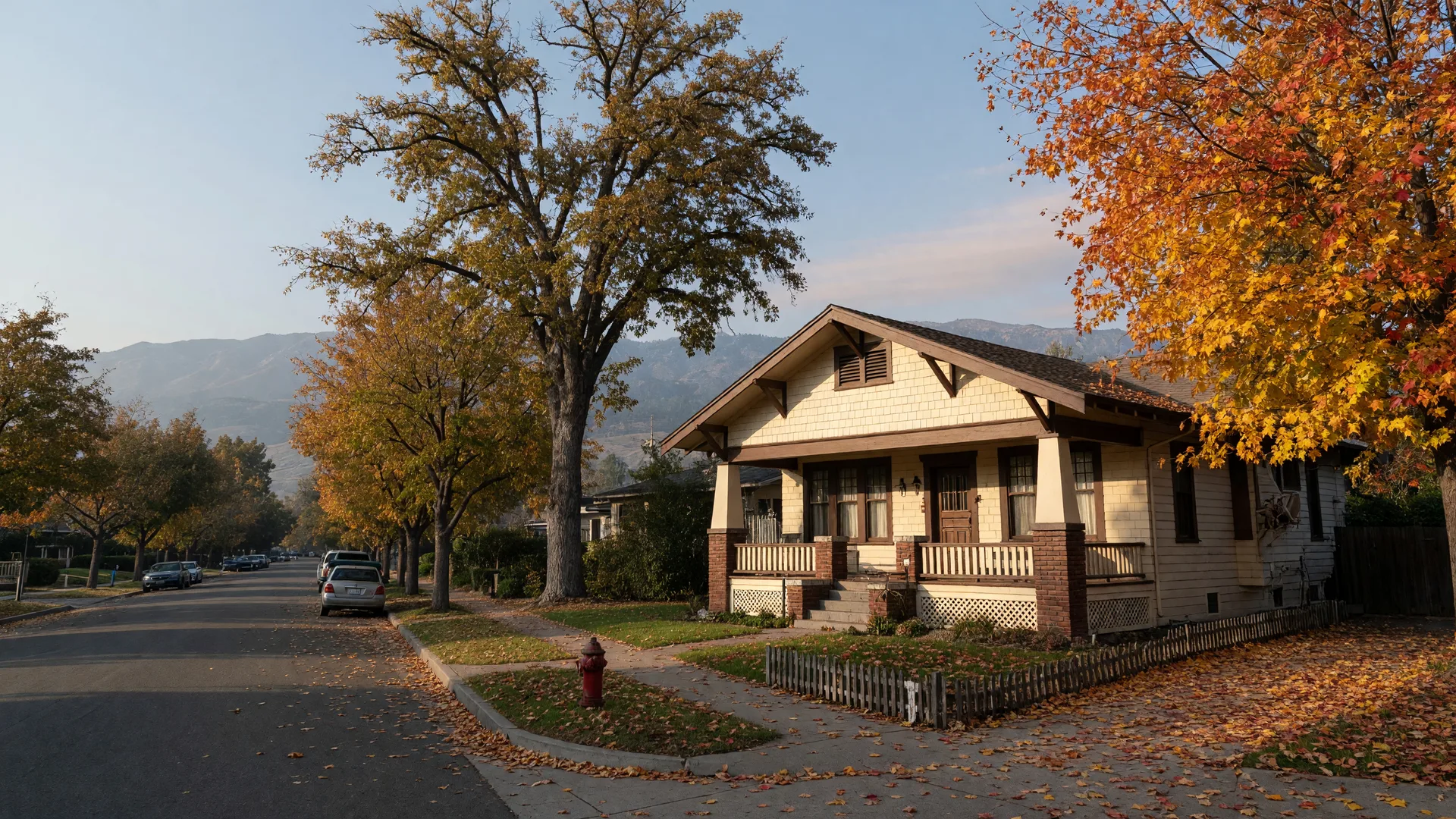 A cream Craftsman bungalow with a hipped gable roof and a wide front porch on a tree-lined street in Merced's Courthouse Park neighborhood on a late-October morning, golden liquidambar leaves on the sidewalk and the hazy Sierra Nevada foothills on the horizon