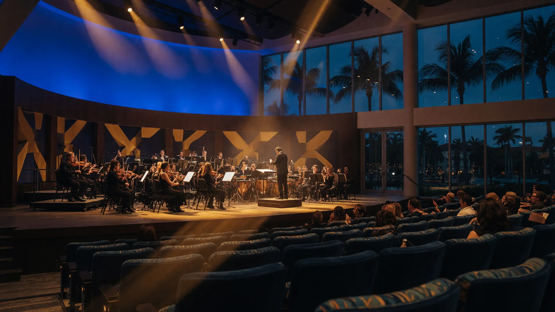 A concert in a modern performing arts center with a band on a warmly lit stage in deep blue and gold light and the audience softly blurred in the foreground