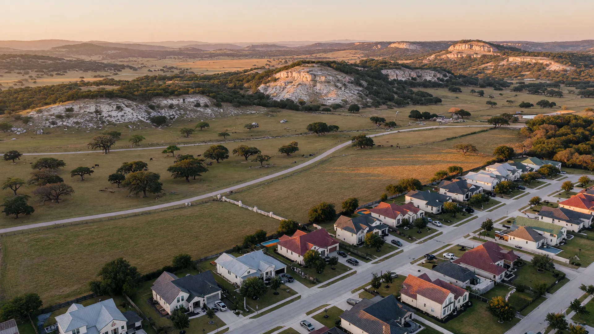 Aerial view of a Texas Hill Country valley at golden hour showing a master-planned neighborhood transitioning to open ranch land with live oaks and limestone hills