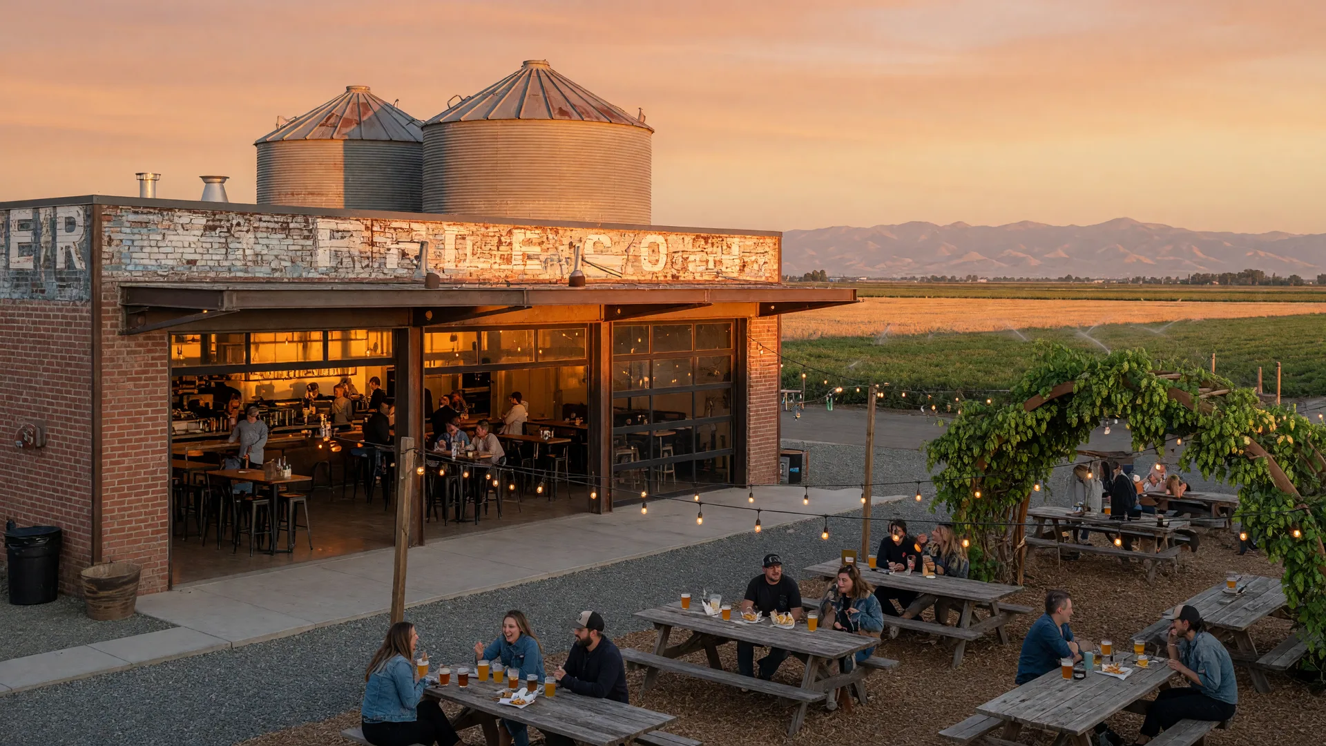 The Dust Bowl Brewing Co. brewery taproom at 3000 Fulkerth Road in Turlock at golden hour, a brick and steel building with glowing glass garage doors, string lights over a beer garden with wooden picnic tables, and the Sierra Nevada foothills on the horizon