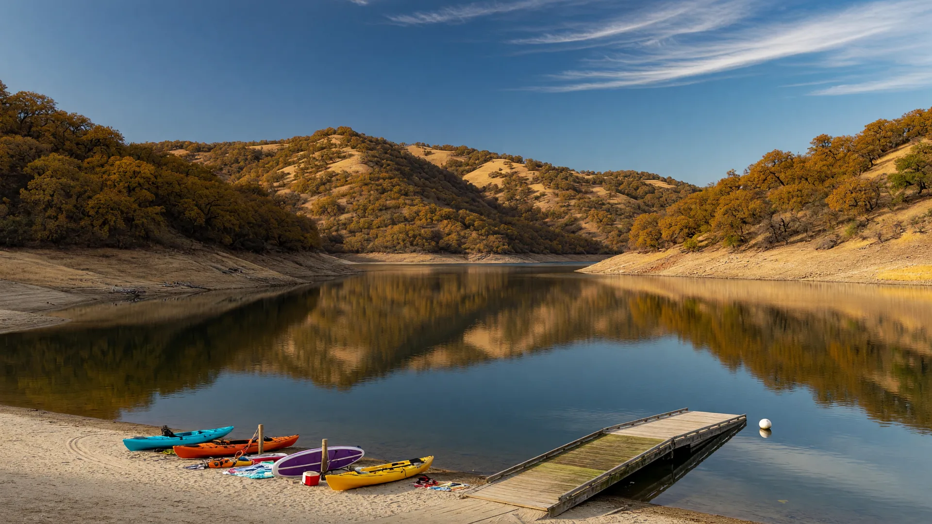 A calm Sierra-foothill reservoir in the Central Valley on a clear October afternoon, golden oak-covered hillsides, a sandy swim beach with colorful kayaks and paddleboards pulled up on the sand, glassy water reflecting the amber hills, and a wooden boat launch ramp