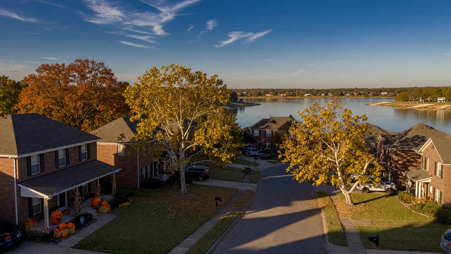 A late-October street in a North Texas lake community with turning trees, pumpkins on front porches, and a reservoir beyond the rooftops