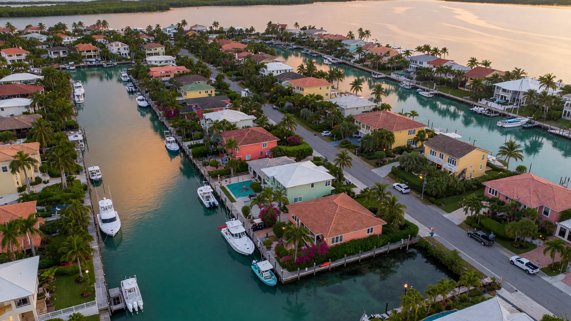 Aerial view of a Florida Keys waterfront neighborhood at golden hour with turquoise canals lined by luxury waterfront homes, private docks with boats, and palm trees