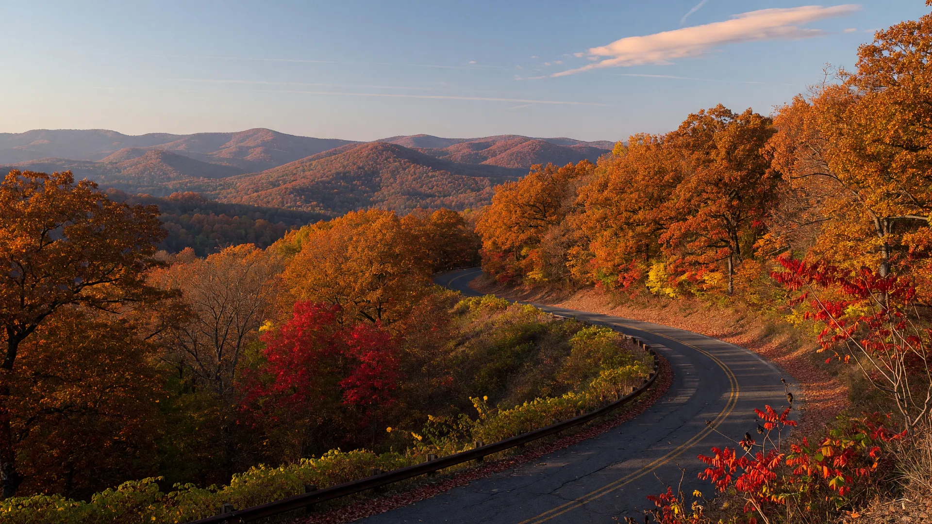 A two-lane scenic road winding along an Oklahoma ridgetop in peak autumn, with oak and maple trees blazing in amber, orange, and gold under late-afternoon light