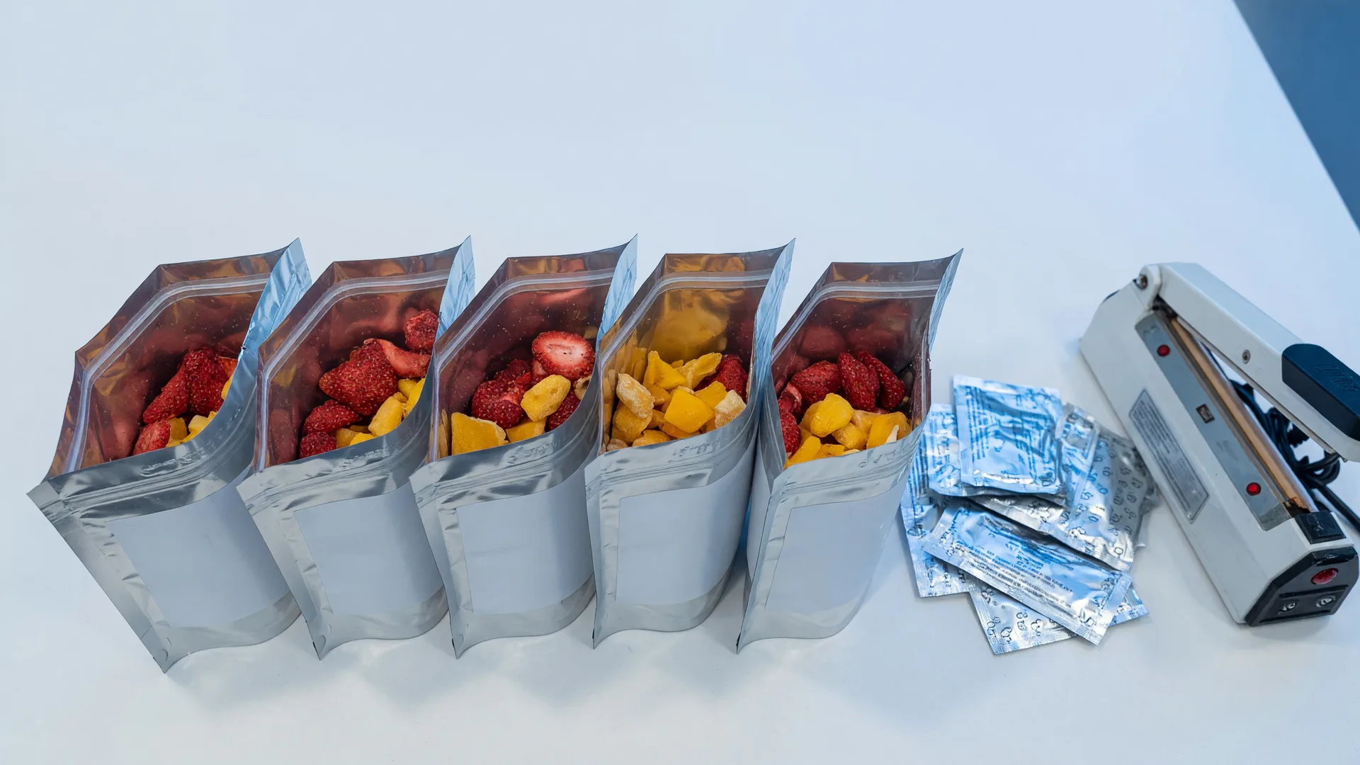 Mylar pouches filled with freeze-dried strawberriesand mango next to oxygen absorber sachets and a heat sealer