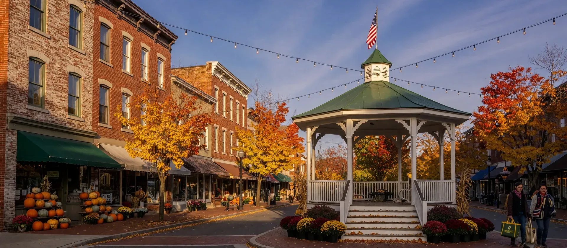 A small Pennsylvania borough main street in mid-October, a town circle with a bandstand, maples in full gold and crimson, pumpkins and mums outside brick storefronts, string lights overhead and a few pedestrians with market bags in warm late-afternoon light
