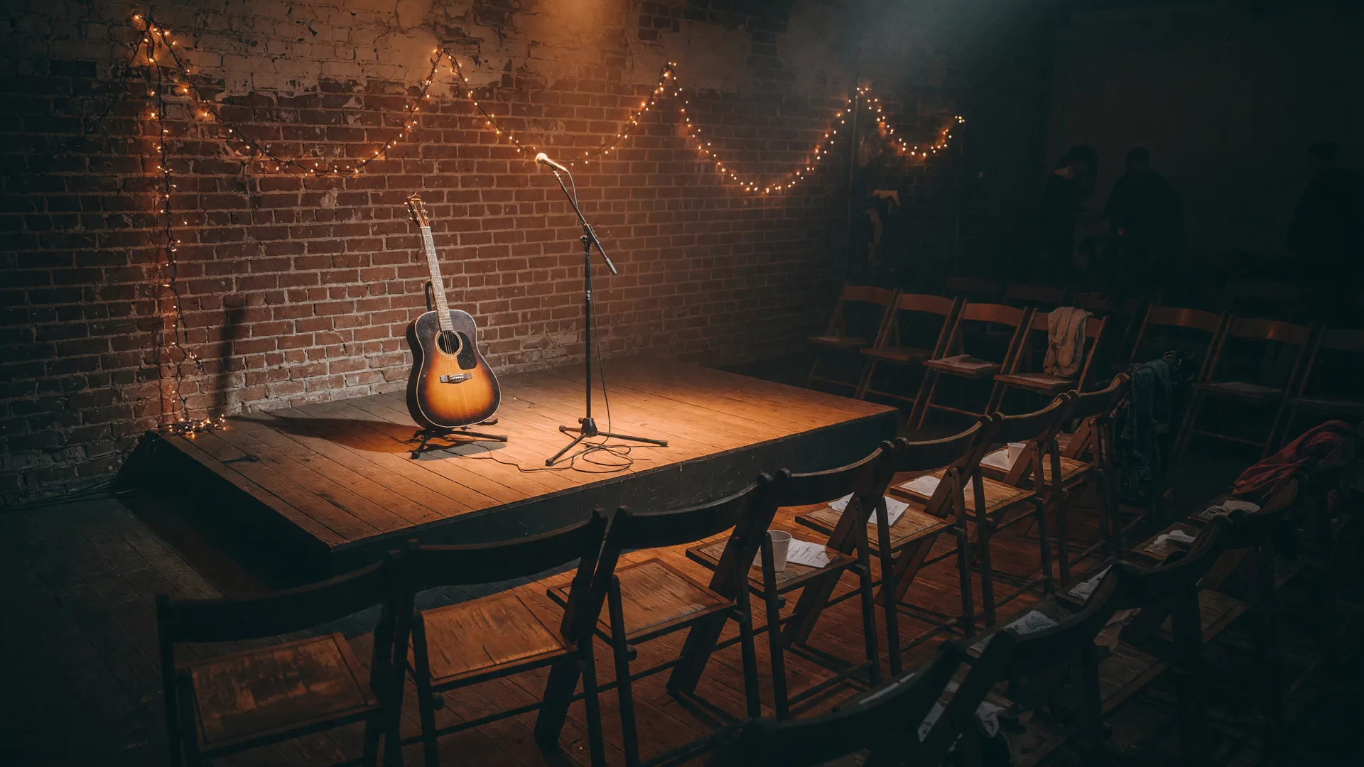A moody small-room stage with a microphone and acoustic guitar under warm string lights in a Pasadena listening room