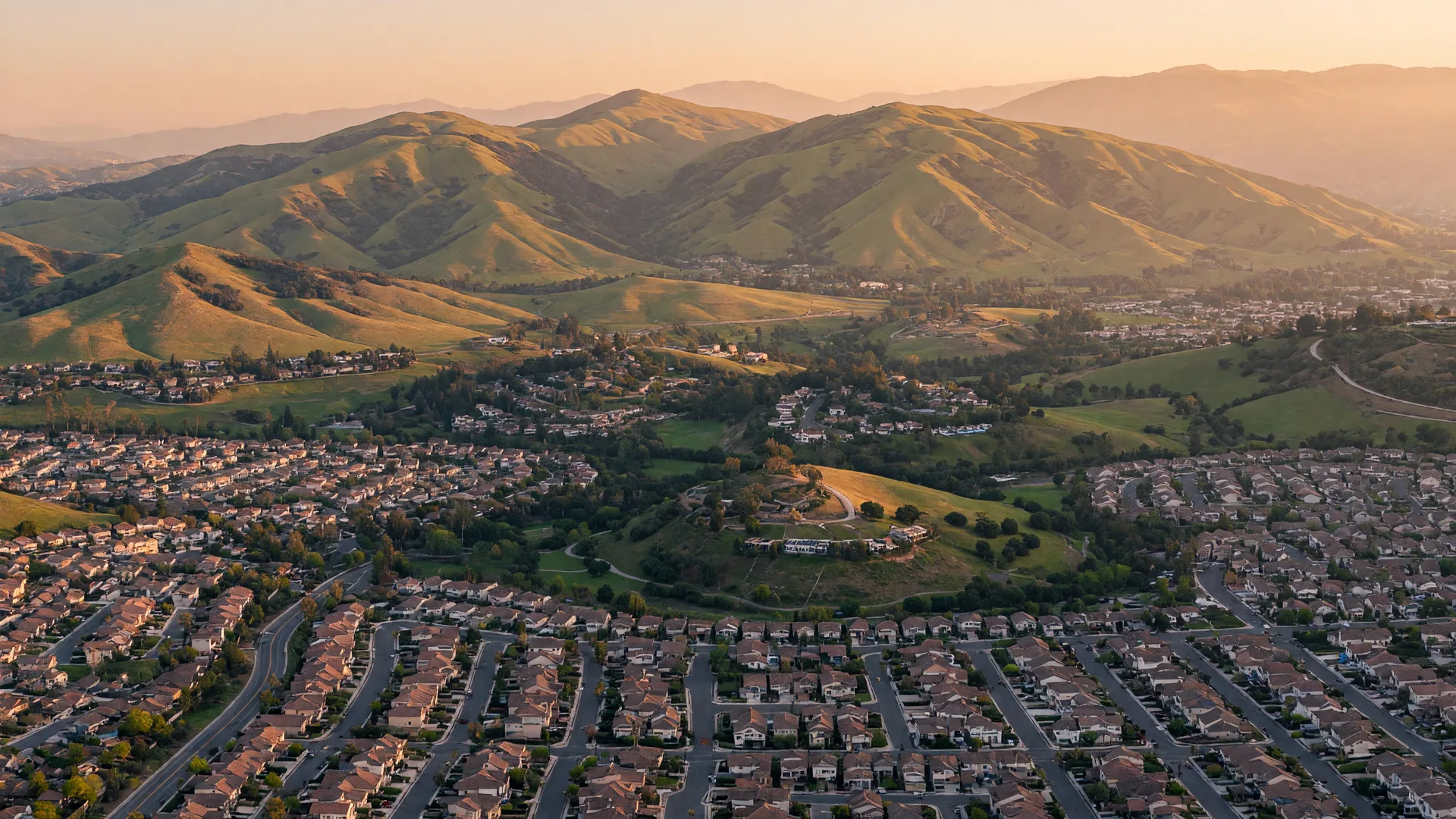 A wide aerial view of Santa Clarita Valley neighborhoods at golden hour, with rooftops nestled against green mountains
