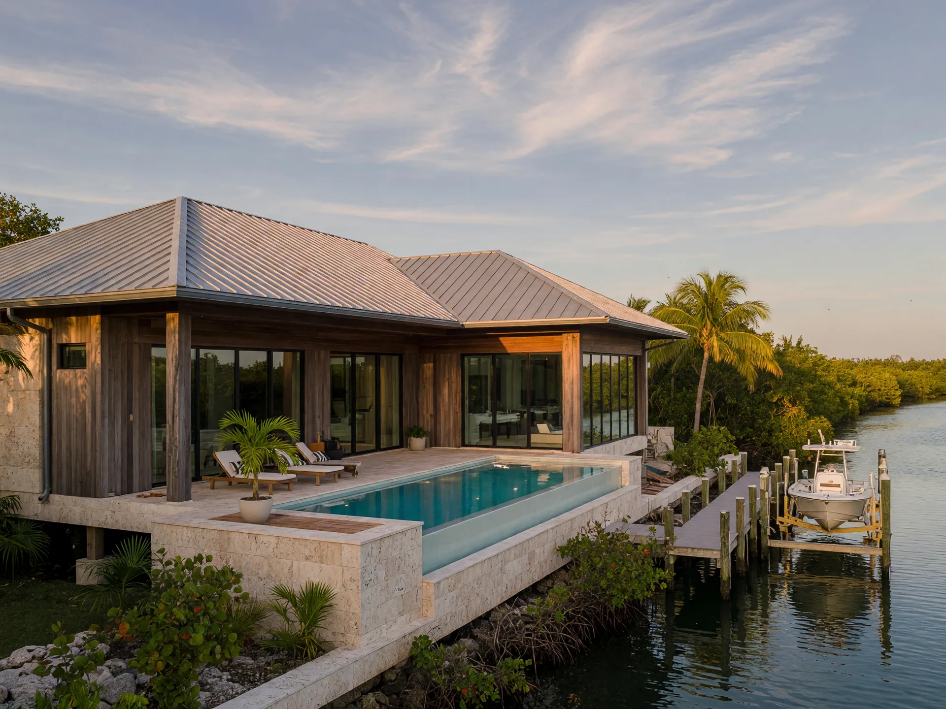 A modern elevated coastal waterfront home in the Florida Keys in late-afternoon golden light, standing-seam metal roof, deep horizontal overhangs, natural limestone and cypress cladding, floor-to-ceiling impact glass opening onto a resort-style pool terrace, a private dock and boat lift on a deep-water canal