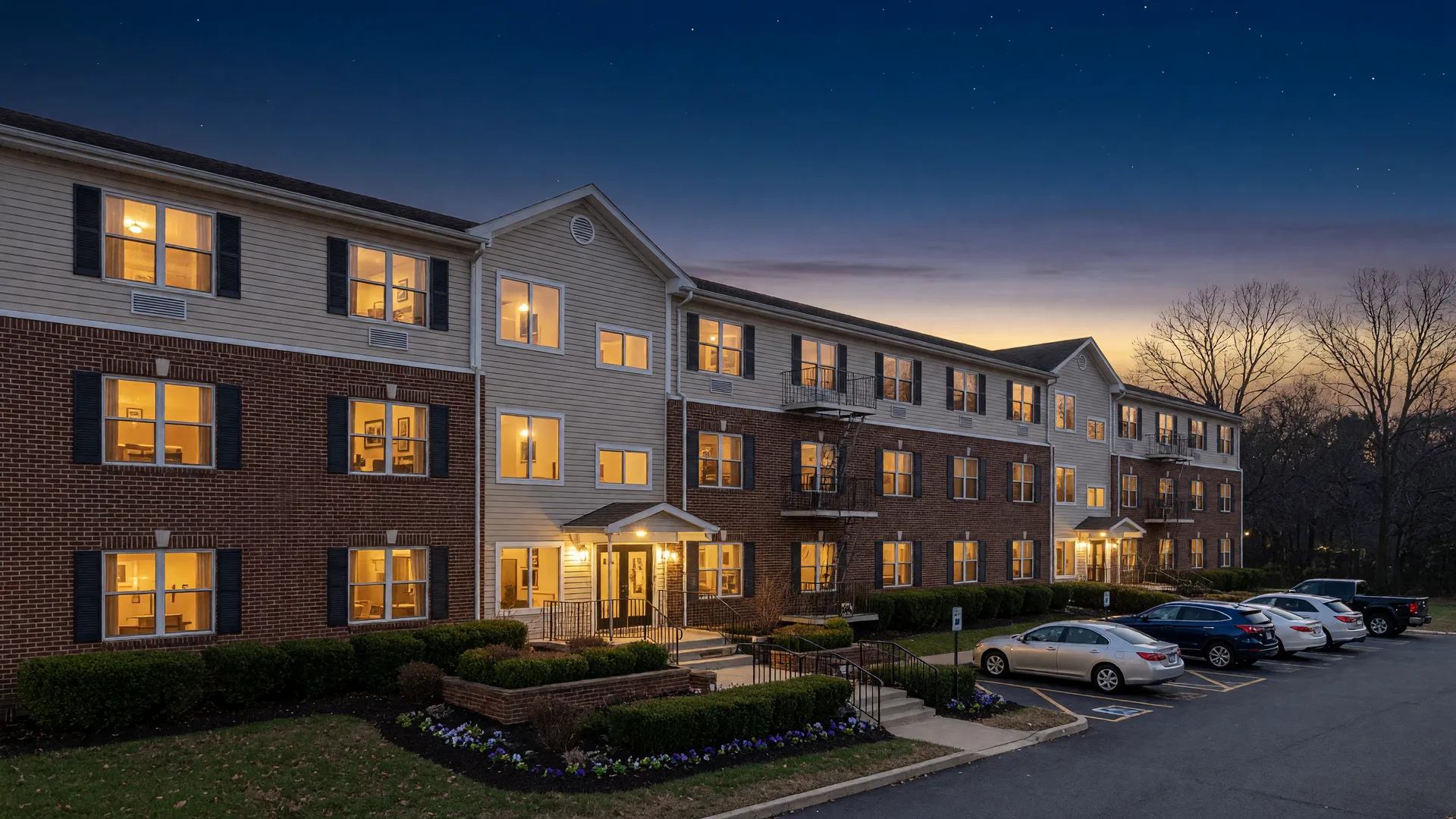 A three-story multifamily apartment building glowing with warm window light at blue hour under a deep navy dusk sky