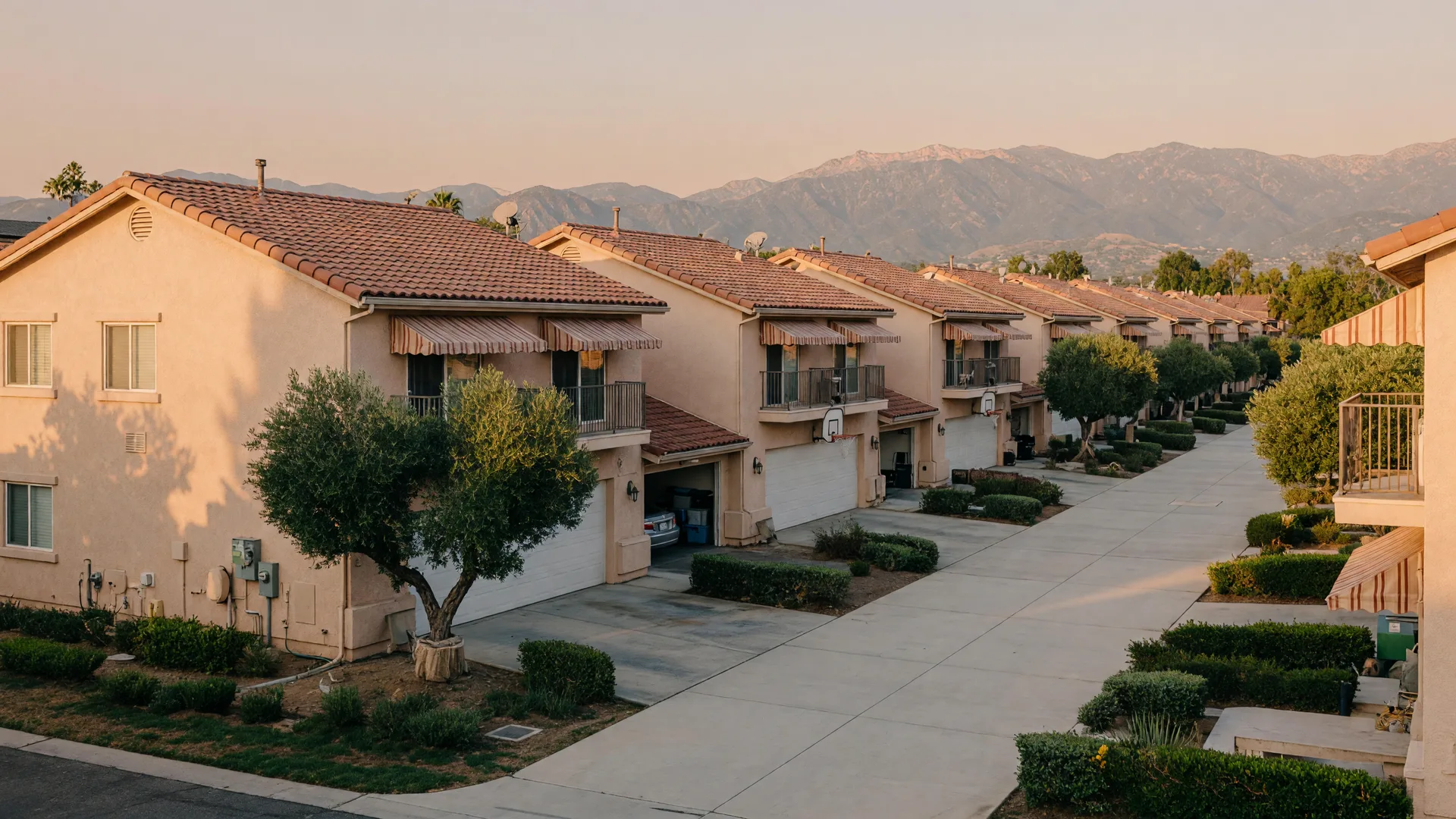 A quiet Newhall condo community at golden hour: two-story stucco buildings with red tile roofs, tree-lined walks, and hazy mountains beyond