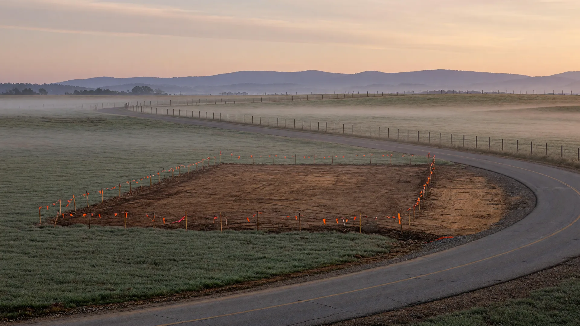 Rural North Georgia county road at dawn bending past pasture with a freshly graded, stake-and-flag marked homesite and blue ridge foothills beyond