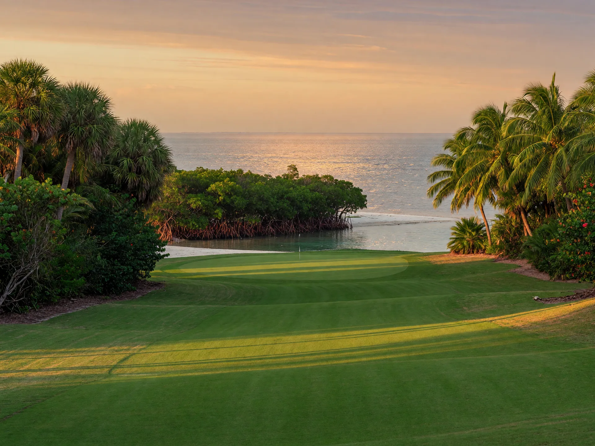 A championship golf fairway in a tropical Florida Keys island club at late afternoon, manicured green grass framed by dense palms and native coastal foliage, with a calm ocean channel and white sand visible beyond a distant stand of mangroves