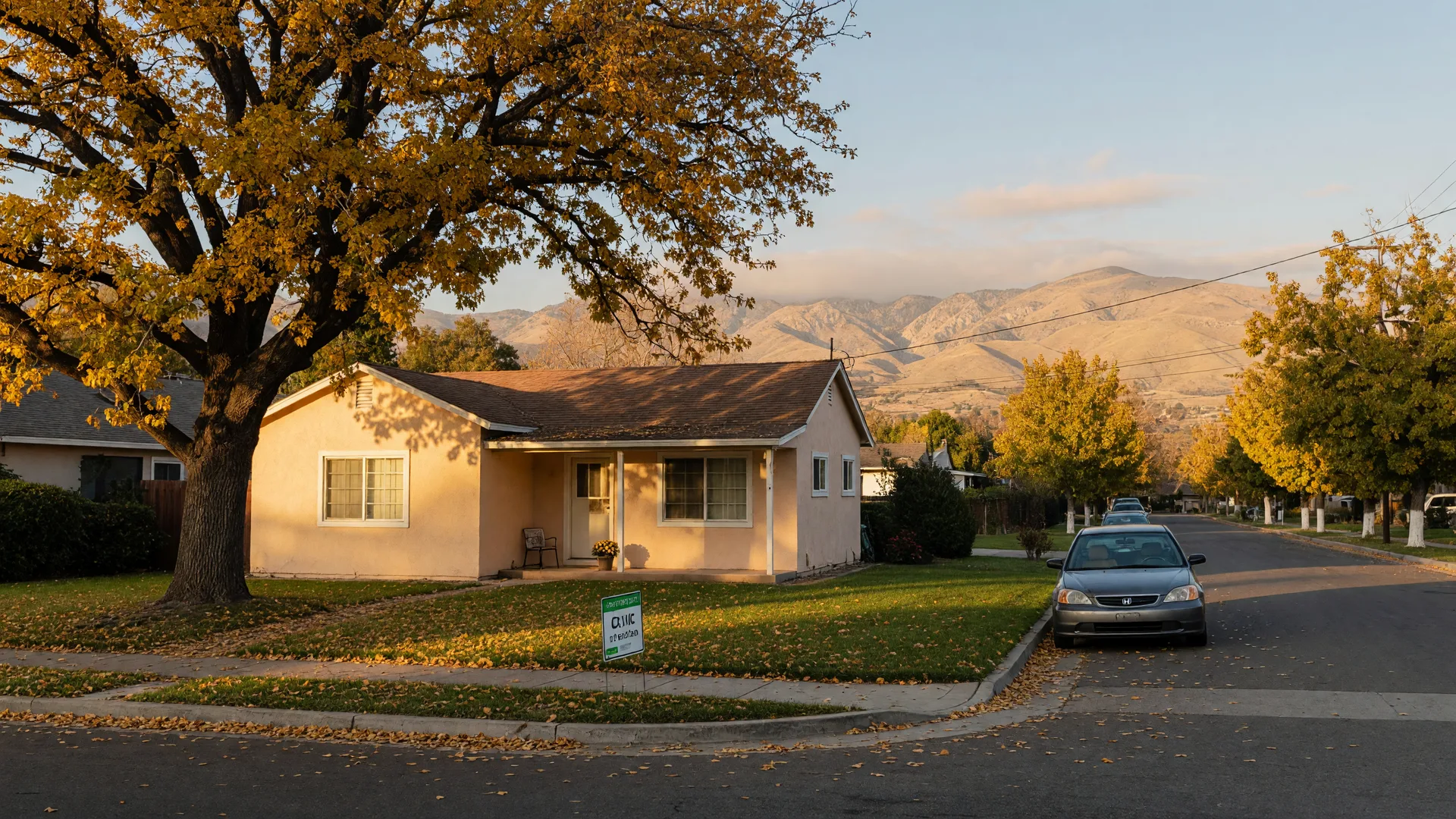 A modest single-story starter home in a Merced County neighborhood on a mid-October afternoon, with a neat lawn, a small yard sign, golden leaves falling from a valley oak, and the Sierra Nevada foothills on the horizon