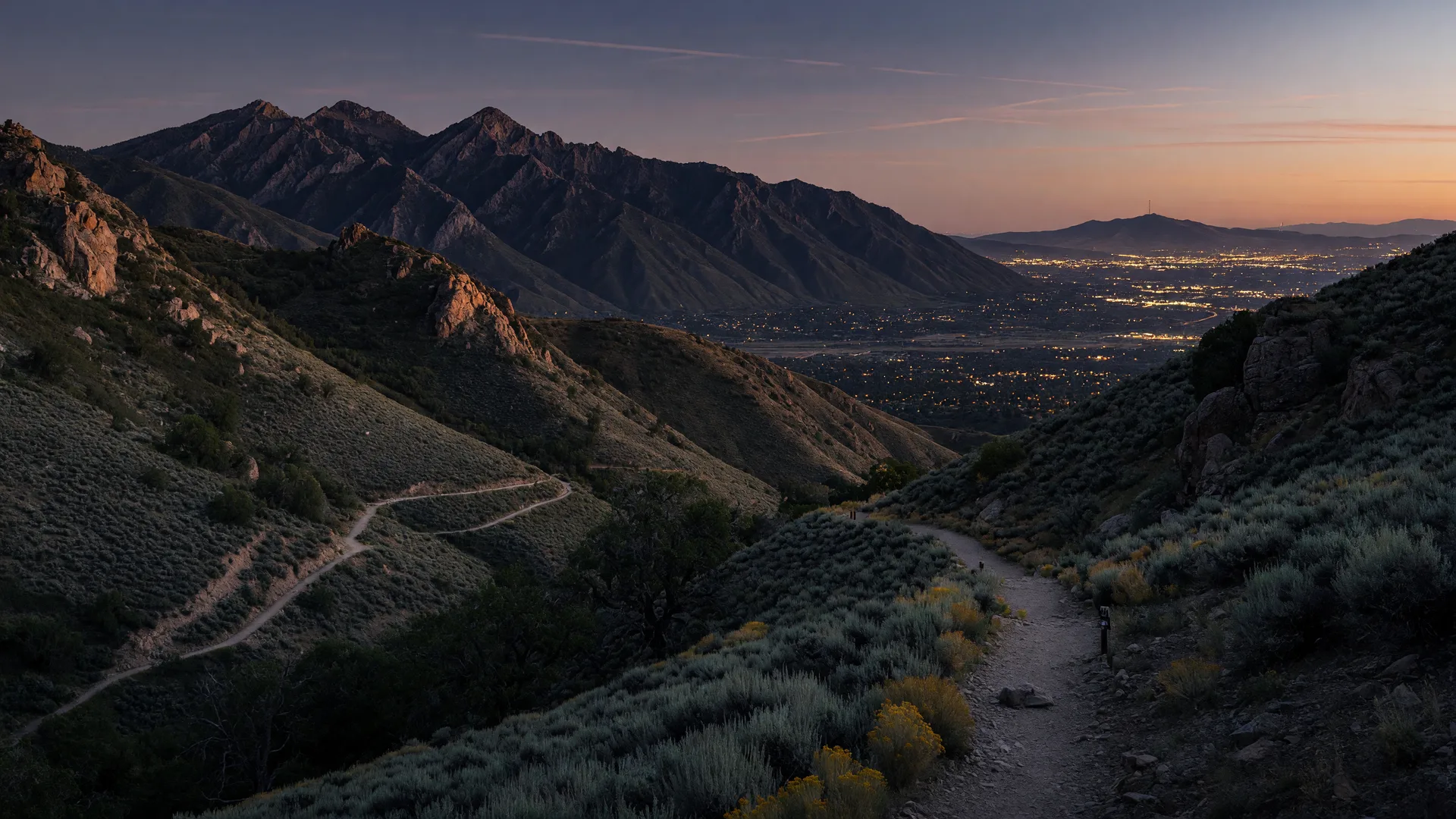 Dusk view of the Wasatch foothills on the Draper and Sandy border, with dark sage-covered slopes in the foreground and Salt Lake Valley city lights glowing below the ridgeline