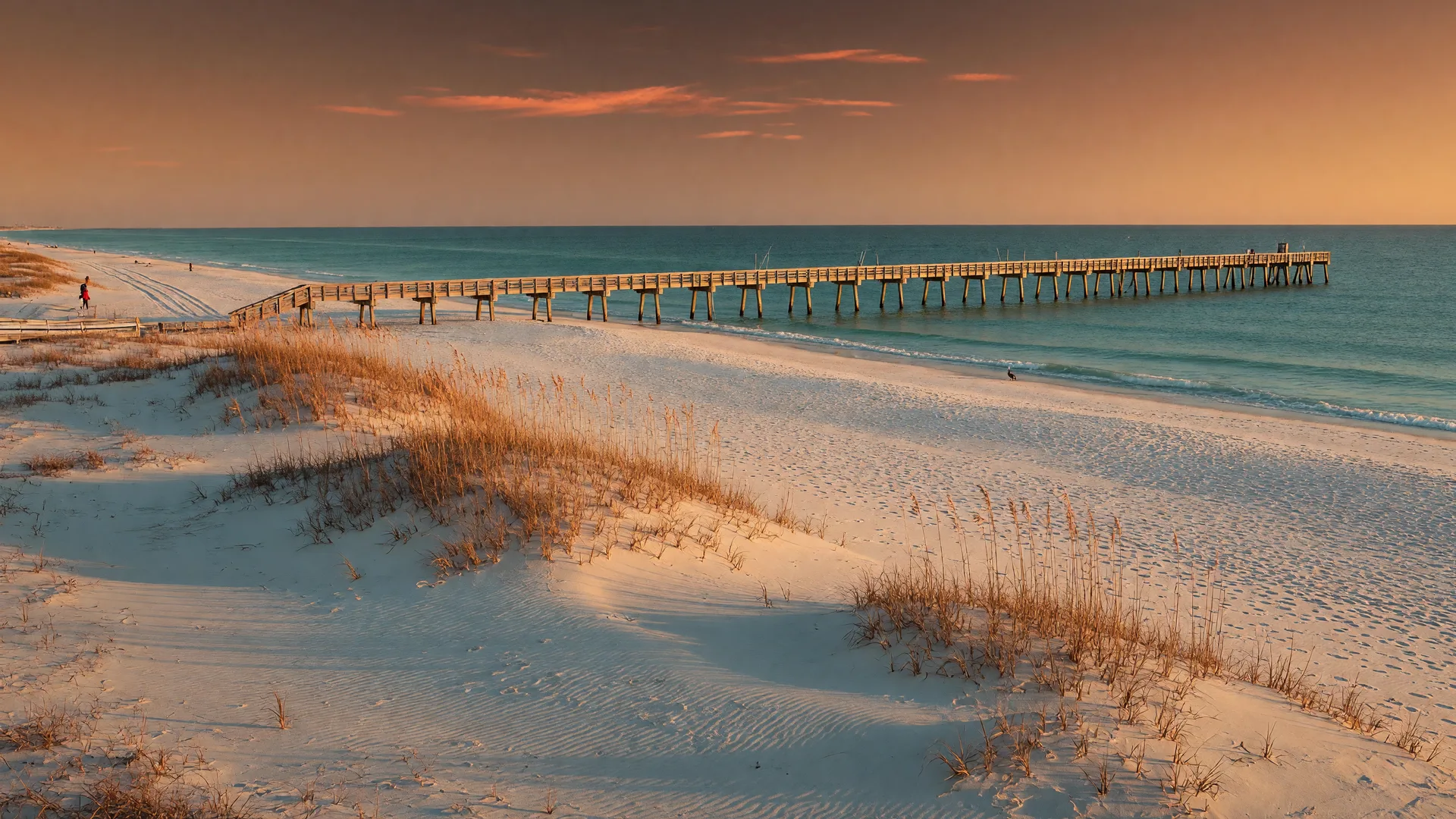 An empty stretch of Pensacola Beach at late-autumn golden hour with wind-rippled sugar-white sand, sea-oats dunes, calm turquoise Gulf rollers, and a long wooden pier under a pink and amber sky.