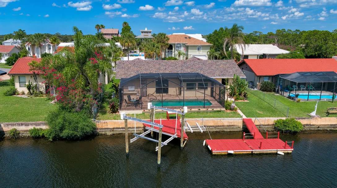 A waterfront Port Richey home at golden hour viewed from the canal, with a concrete seawall, a private wooden dock with a moored boat and boat lift, and palm trees framing the scene