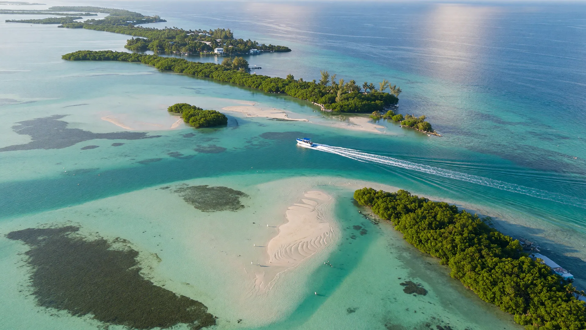 Aerial view of the Upper Florida Keys at mid-morning, a chain of lush green islands surrounded by crystal-clear turquoise shallows with a small boat crossing between islands and sandbars visible beneath the surface