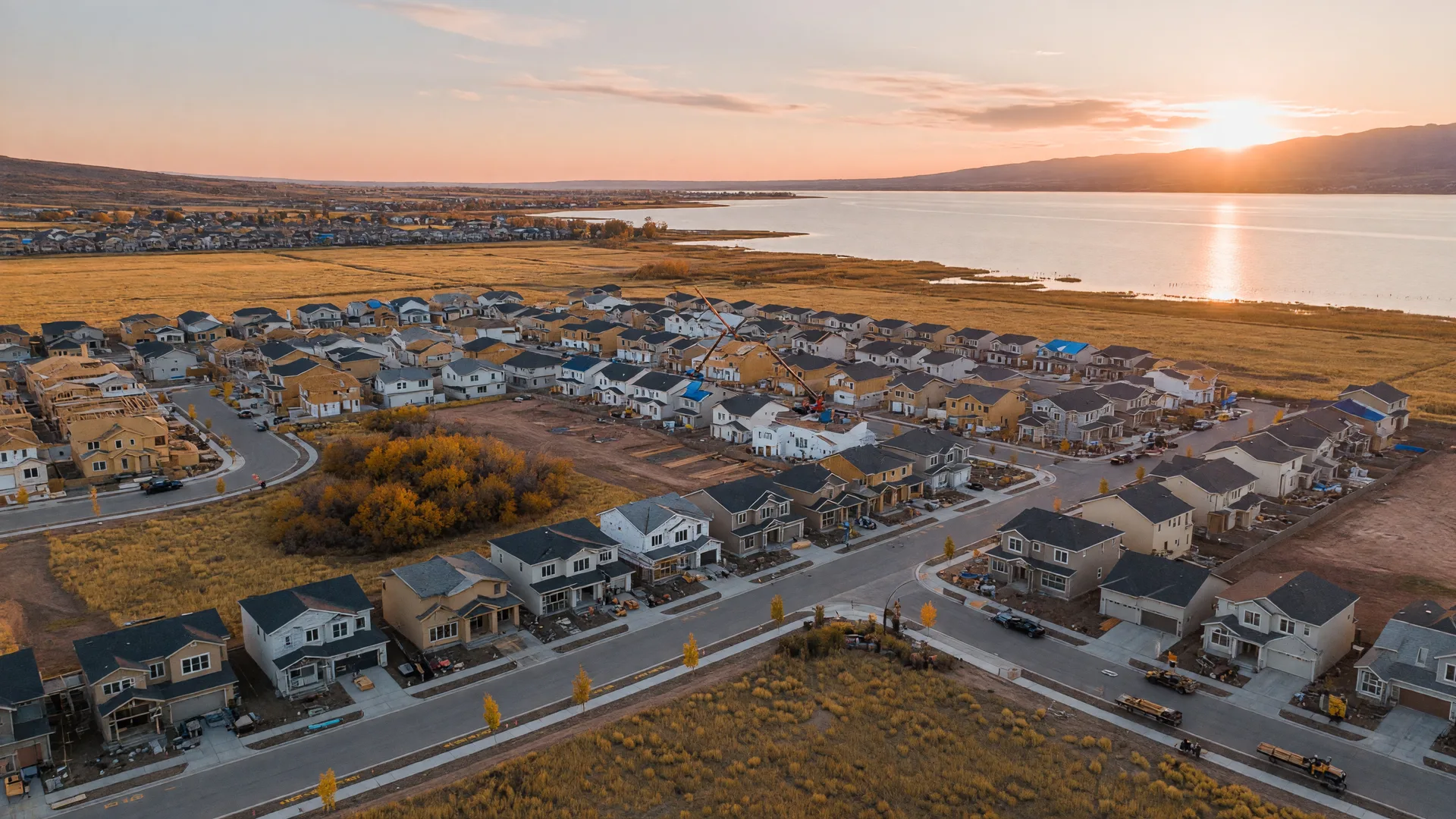 Aerial view of an active new-construction master-planned community in Utah County with homes in framing and finishing stages, cranes, and Utah Lake in the distance