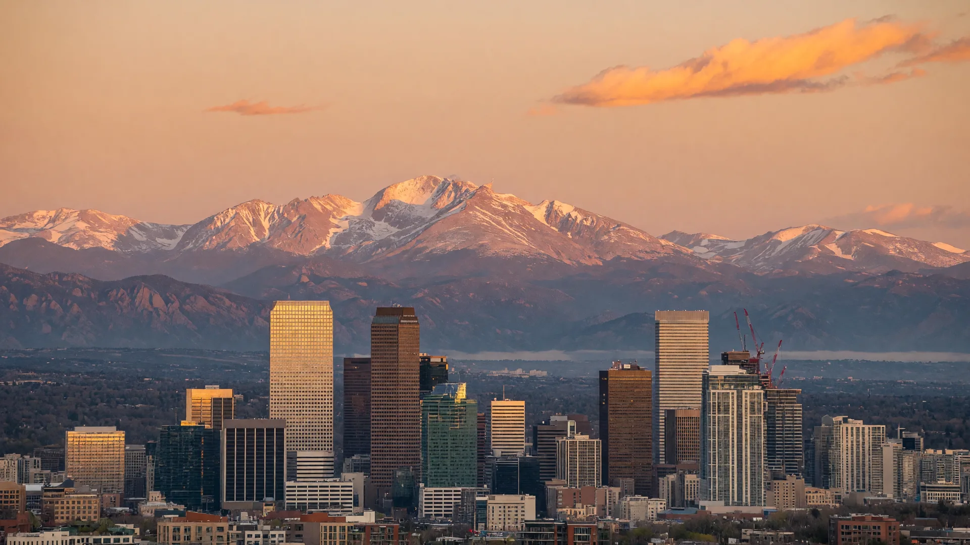 A panoramic view of the Denver, Colorado skyline at golden hour with the Rocky Mountains rising behind it