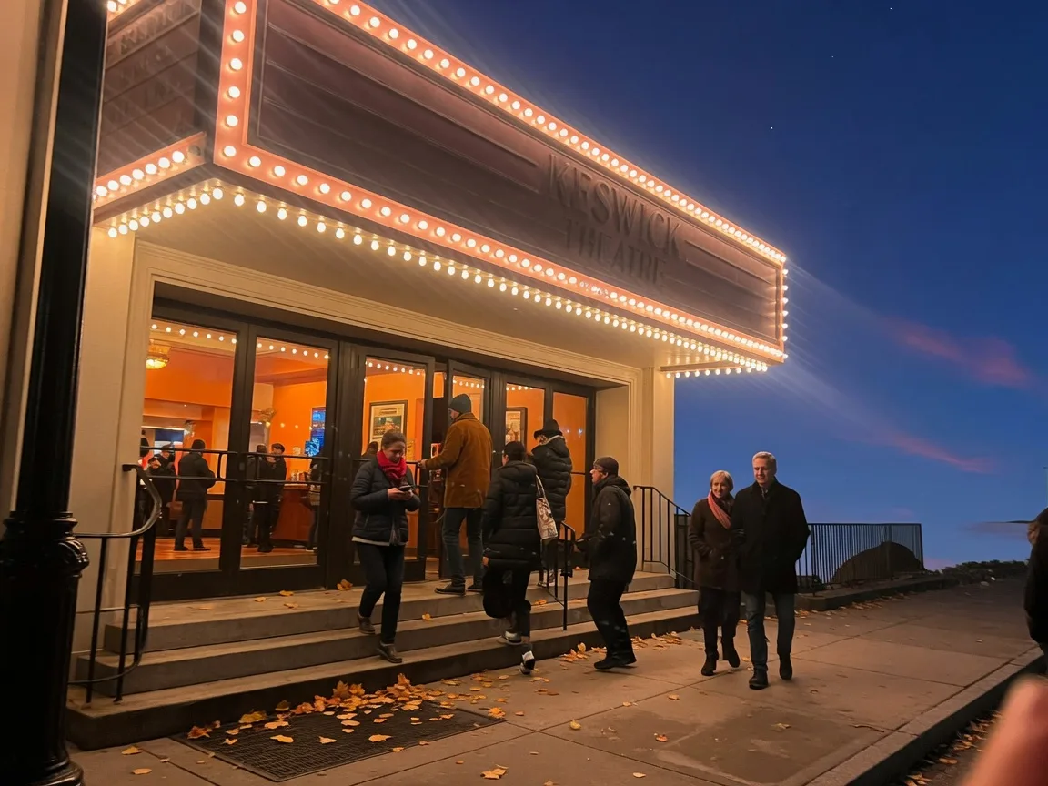 The Keswick Theatre entrance at dusk on a late-October evening, the historic marquee lit warm against a deep blue sky, golden leaves on the sidewalk, a small crowd of adults in jackets heading toward the glowing lobby doors