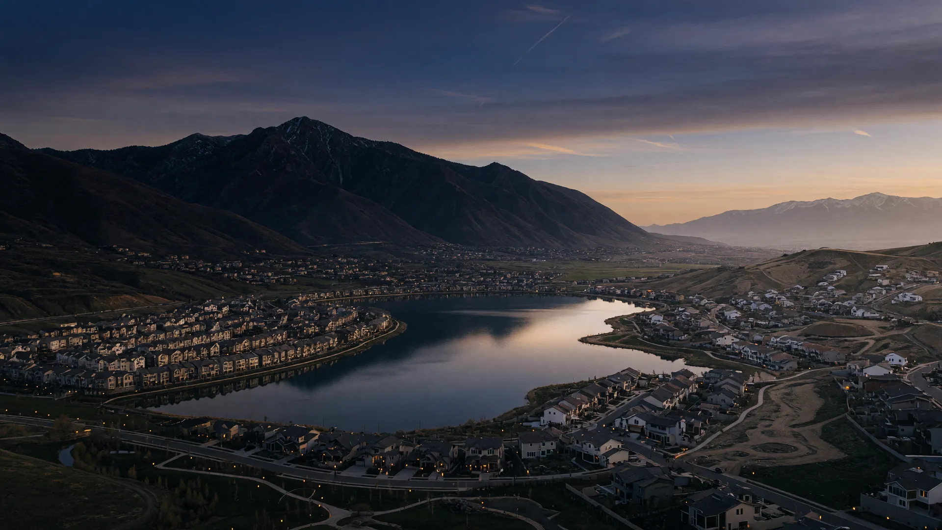 Aerial dusk view of Utah's southwest Salt Lake Valley with a master-planned community around a lake and foothill subdivisions beneath the Oquirrh Mountains
