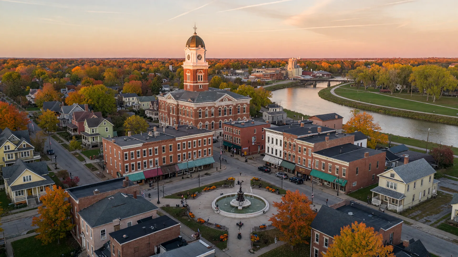 A wide aerial view of Troy, Ohio in mid-October, looking down over the historic public square with its central fountain and preserved 19th-century brick storefronts, the red-brick Miami County Courthouse dome rising above the roofline, tree-lined streets of Victorian homes with maples turning orange and gold, and the Great Miami River curving through the city in the distance, in warm late-afternoon light