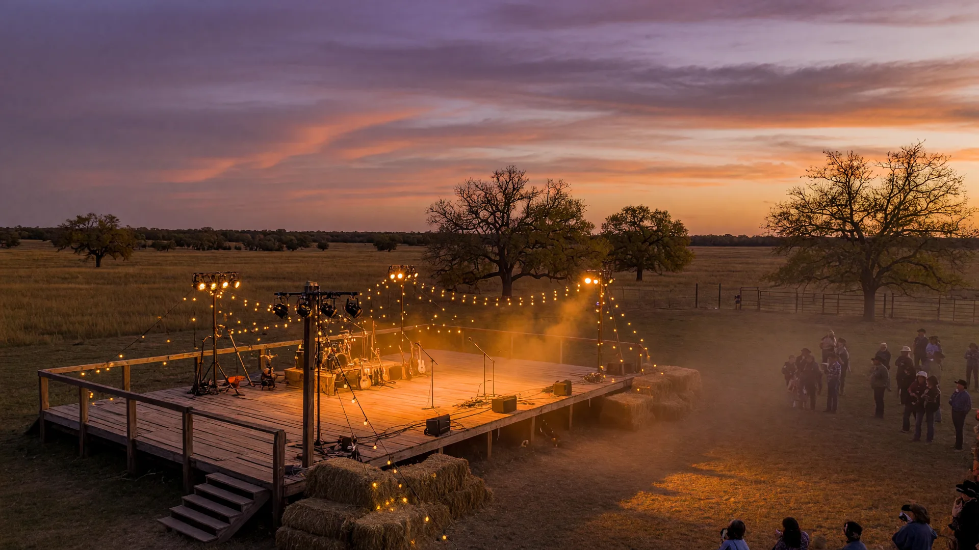 An outdoor country music festival stage on a Texas ranch at dusk, rustic wooden stage under warm amber floodlights with string lights, hay bales, and a purple-orange sunset sky
