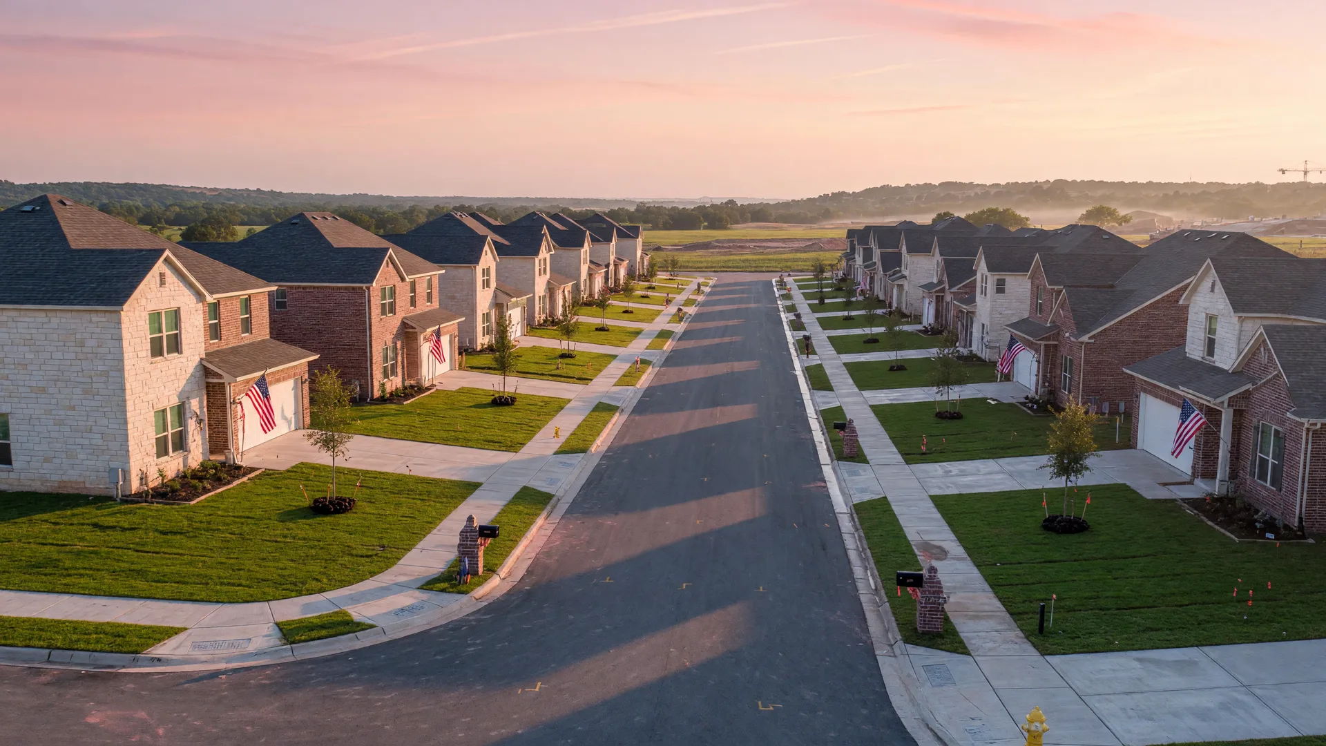 A new-construction suburban street in the Texas Hill Country at sunrise with flags on the porches