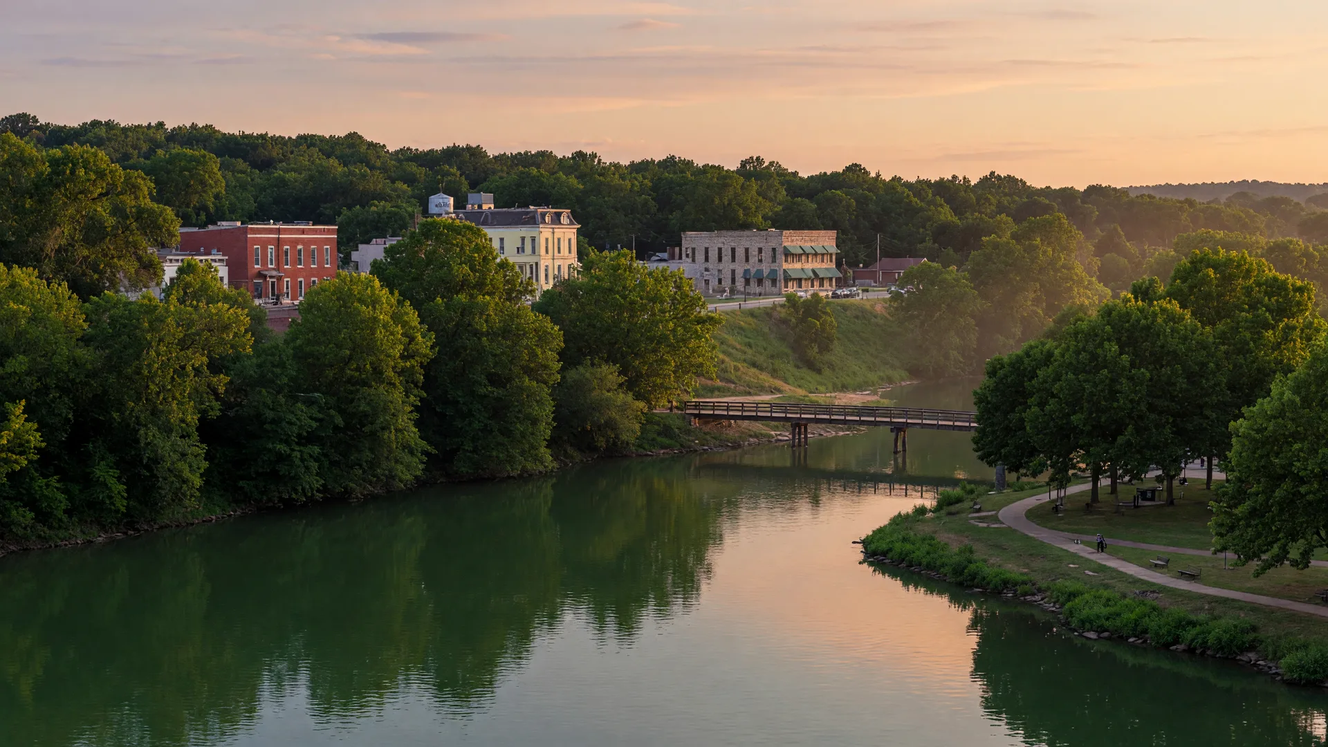 The Finley River flowing through Ozark, Missouri at golden hour, with a riverside walking path, wooden footbridge, and tree-lined banks