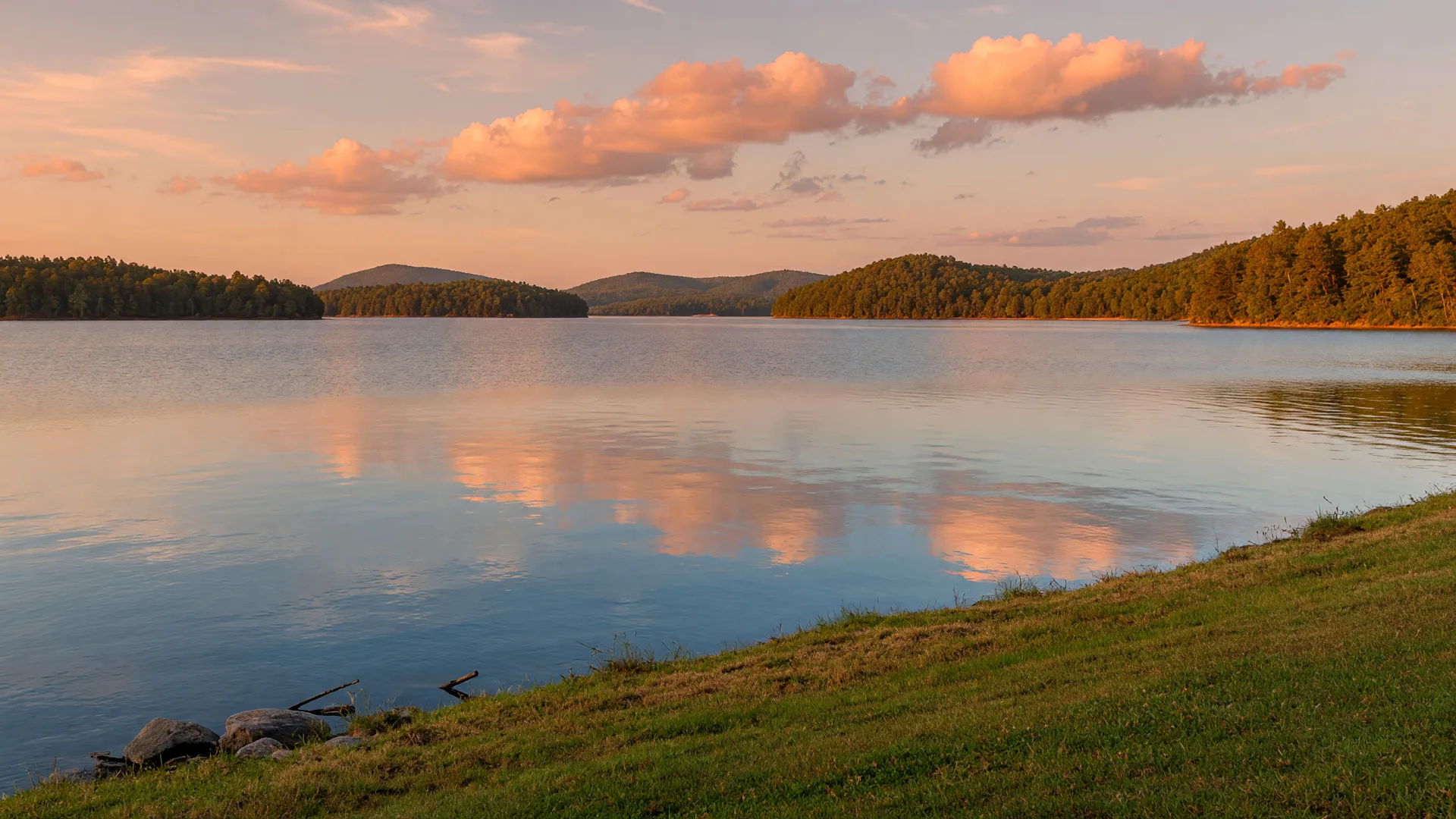 Lake Hartwell at golden hour with calm water, pink and orange clouds reflected on the surface, and tree-covered hills on the horizon