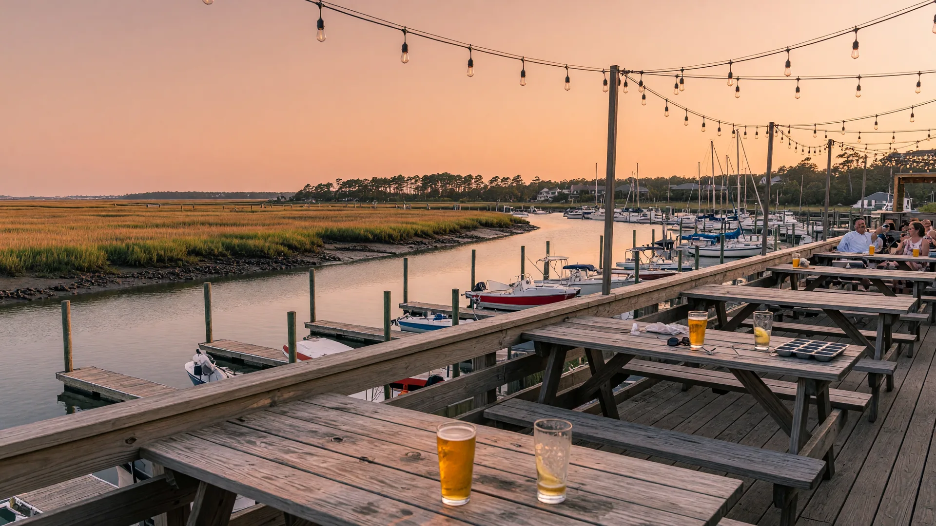 Waterfront brewery patio at golden hour with wooden tables, glasses of golden lager catching the low sun, string lights overhead, and sailboats bobbing in the marina behind